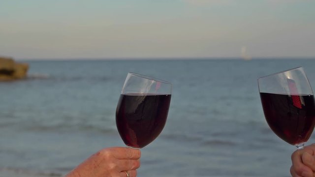 Toast mit zwei Rotweingl&auml;sern am Strand. Beim Ansto&szlig;en in Zeitlupe schwappt der Wein &uuml;ber.