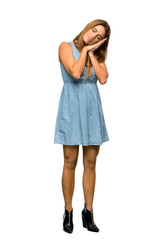 A full-length shot of a Blonde woman with jean dress making sleep gesture in dorable expression over isolated white background