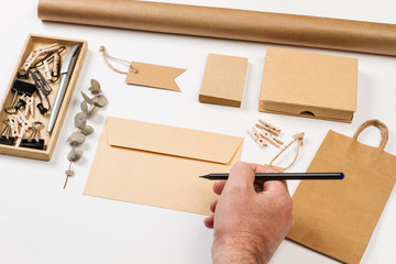 Mano de hombre con un bolígrafo y un sobre, tarjetas, caja, etiquetas y papel kraft marrón sobre...