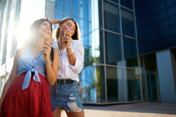 Two young female friends having fun and eating ice cream. Cheerful caucasian women eating icecream outdoors in the city. Pretty girls posing with ice-cream. Summer time concept.