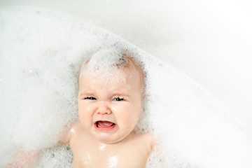 A crying Baby girl bathes in a bath with foam and soap bubbles