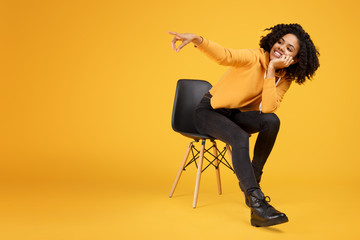 Charming african american young woman with beautiful smile dressed in casual clothes sitting on the stylish chair and point finger away over yellow background.