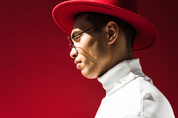 side view of stylish mixed race man in hat and glasses posing isolated on red with copy space