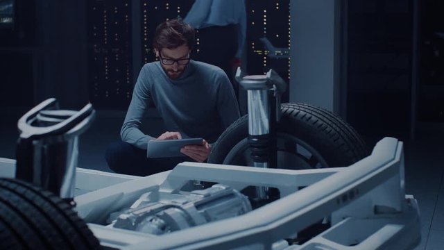 Automobile Design Engineer Sitting Beside Hybrid Electric Car Chassis Platform Prototype, Using Tablet Computer for Design Enhancement. Facility with Vehicle Frame with Suspension, Wheels, Engine.
