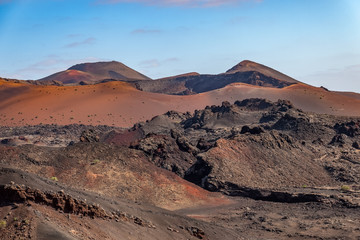 A volcanic landscape with peaks, craters, and orange sand. There are small bushes scattered around, and blue sky in the background. Taken in the Timanfaya National Park, Lanzarote.