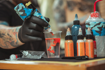 Red tattoo ink spreading in glass of water on the foreground, tattoo artist working with a...