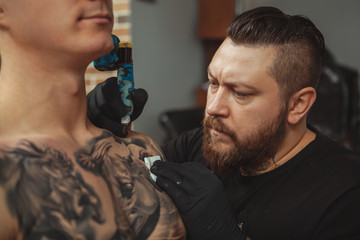 Close up of a concentrated bearded tattoo artist working, tattooing shoulder of a male client....