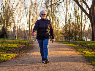 Fototapeta premium A senior woman with white hair listening to music with headphones walking in an urban park