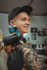 Vertical close up of a handsome young man smiling, getting tattooed on his arm. Cropped shot of hands of professional tattoo artist working, making a tattoo on arm of a client