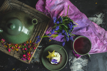 on a decorated with flower petals and flowers a dark wooden table, stands a kettle with tea, a cup and a chocolate cupcake