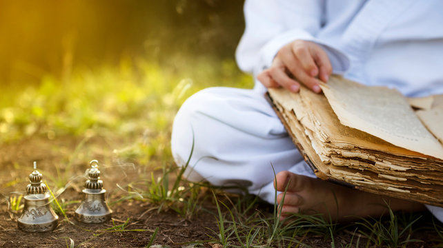Buddhist  monk sits in  yoga lotus position meditating, reading old open book of wisdom, and there are a number  Tibetan bells and burn incense at meadow. beautiful sunset background