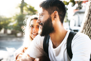 Fototapeta premium Close up portrait of confident bearded man looking away smiling while is embraced by his girlfriend which is looking at him outside while dating in the city.