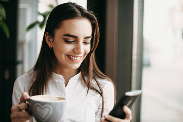 Attractive caucasian woman using a smartphone smiling while holding a cup of coffee near a window in a coffee shop.