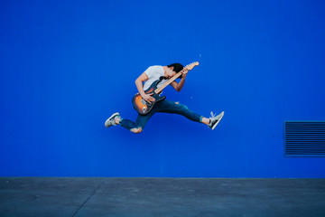 Plakat young man jumping with electric guitar on blue background