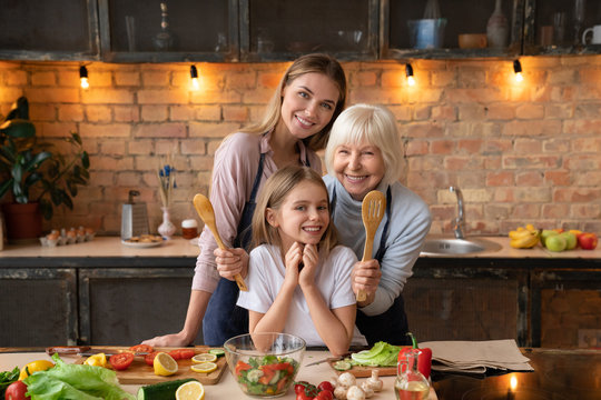 Horizontal Front View Shot Of Happy Family That Lookin In Camera While Cooking In Kitchen. Grandmother Hug Her Granddaughter With Mother Behind Back