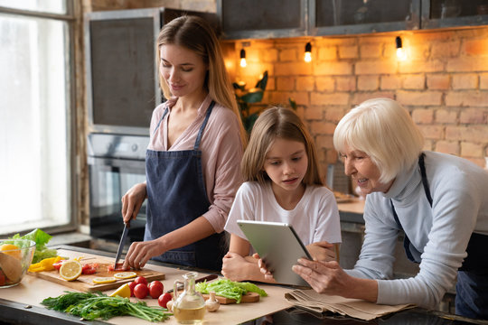 Horizontal Shot Of Senior Woman That Looking In The Tablet With Her Granddaughter And Smiling. Mother Cooking Vegetatian Dinner In Kitchen