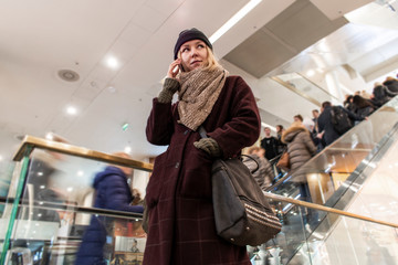 The girl is talking on the phone in a shopping center near the escalator. The flow of people behind the girl, whom she does not notice.