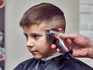 Barber making a haircut to a cute European boy using cutting machine.  Side view. - 259599285