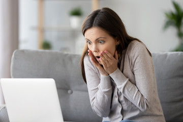 Woman sitting on couch reading message on computer feels shocked