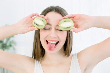 Cheerful beautiful young woman holding two cut kiwis on her eyes for fun joke, smiling and showing tip of her tongue, indoors studio shoot. Beauty and skin care concept