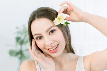 Obraz na płótnie Canvas Beautiful girl with brown eyes and fresh skin posing at light background with flower, skin care concept, beauty spa, bio product. Horizontal