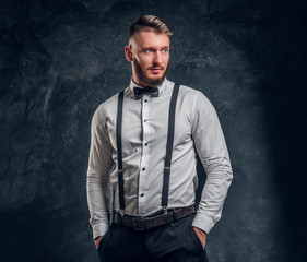 Stylishly dressed young man in shirt with bow tie and suspenders. Studio photo against dark wall background