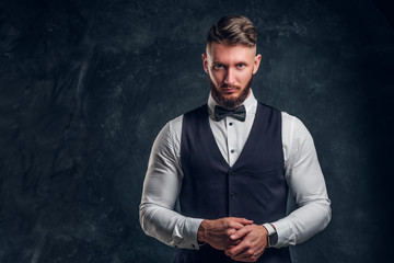 Elegantly dressed bearded hipster in a vest with bow tie. Studio photo against a dark wall background