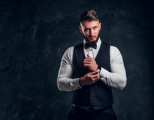 Elegantly dressed bearded hipster in a vest with bow tie. Studio photo against a dark wall background