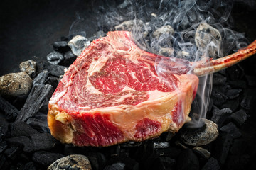 Traditional raw dry aged wagyu tomahawk steak on hot burning charcoal with smoke as closeup on a...