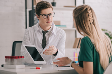 Obraz premium selective focus of handsome doctor in white coat holding digital tablet with blank screen near blonde patient