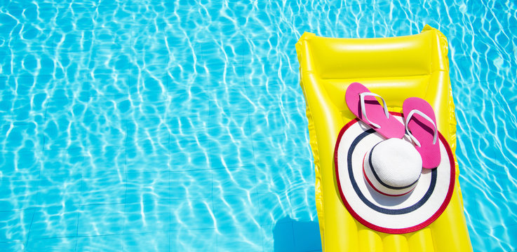 Beach summer holiday background. Inflatable air mattress, flip flops and hat on swimming pool. Yellow lilo and summertime accessories on poolside. Top view and copy space. Banner