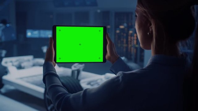 Female Engineer Holds a Tablet Computer with Green Screen Mock Up and Looks at Electric Car Chassis Prototype with Wheels, Batteries and Engine Standing in a High Tech Development Laboratory.