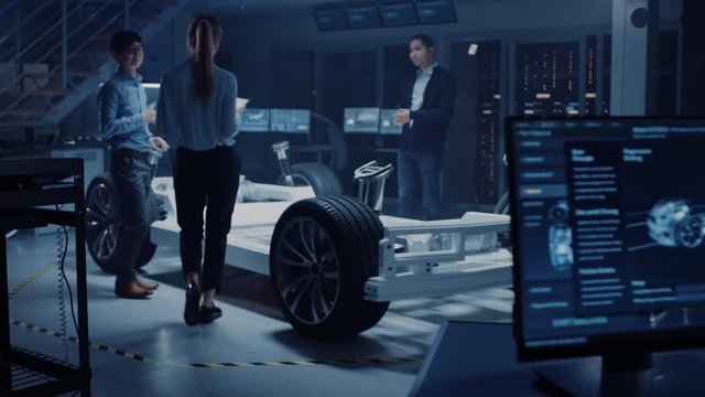 Automotive Design Engineers Talking while Working on Electric Car Chassis Prototype. In Innovation Laboratory Facility Concept Vehicle Frame Includes Wheels, Suspension, Engine and Battery.