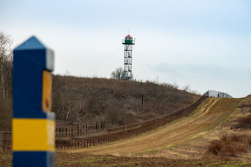 Fototapeta na wymiar Ukrainian-Polish border, barrier fence with a control strip and border pillars. Observation tower in the background. Harsh spring landscape against the blue sky. Defensive security protective concept