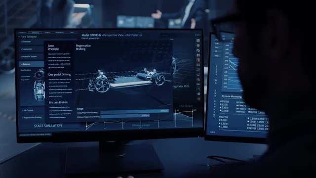 Professional Engineer Works on a Computer with a 3D CAD Software and Tests the Electric Car Chassis Prototype with Wheels, Batteries and Engine Standing in a High Tech Development Laboratory.