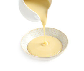 Pouring condensed milk from jug into bowl on white background. Dairy product