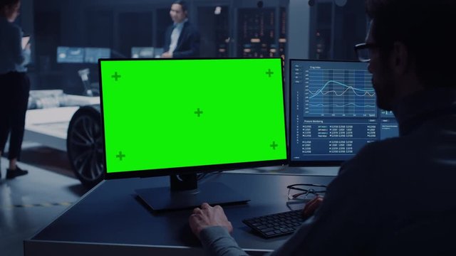 Professional Engineer Works on a Computer with Green Screen Mock Up and Tests the Electric Car Chassis Prototype with Wheels, Batteries and Engine Standing in a High Tech Development Laboratory.