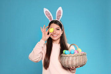 Beautiful woman in bunny ears headband holding basket with Easter eggs on color background - Powered by Adobe