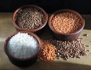 Grains of buckwheat, lentils and rice in clay bowls on a board, against a dark background