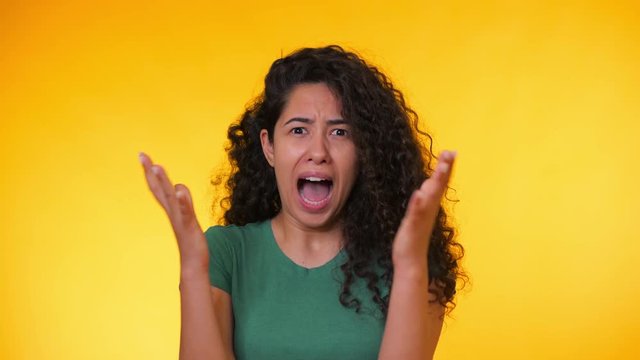 Frightened hispanic woman with curly hair in green t-shirt afraid of something and looks into the camera with big eyes full of horror over yellow background