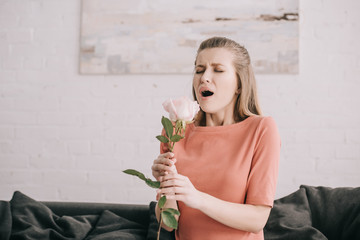 attractive blonde girl with pollen allergy sneezing while holding rose at home
