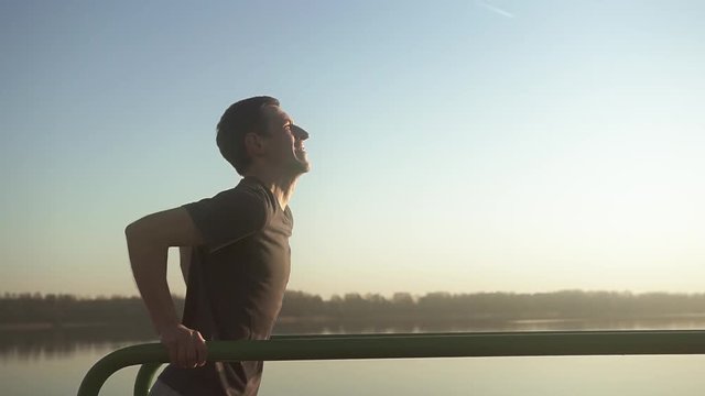 Young thin guy trains on workout bars. Sports activities in the open air on the lake. Slow motion.