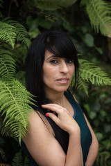 Beautiful mature Latina woman leaning against a green wall of ferns and moss.
