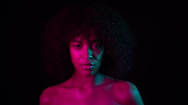 Portrait of young charming african girl in pink neon light at black background. Tempting woman with perfect makeup looking at camera and smiling. Glamour, fashion concept