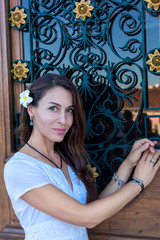 Portrait of beautiful girl with long hair in a white dress and a frangipani plumeria flower on the background of a wooden door with glass and wrought-iron bars