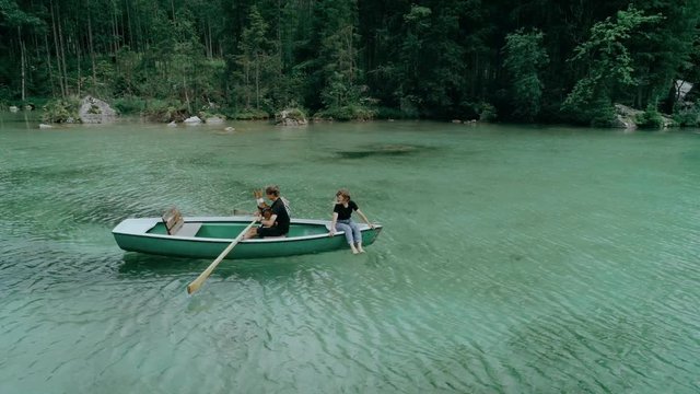 Drone follows small vintage wooden rowing boat with cute millennial couple and their dog enjoying views and nature of beautiful epic mountain lake surrounded by green forest. concept travel blogger