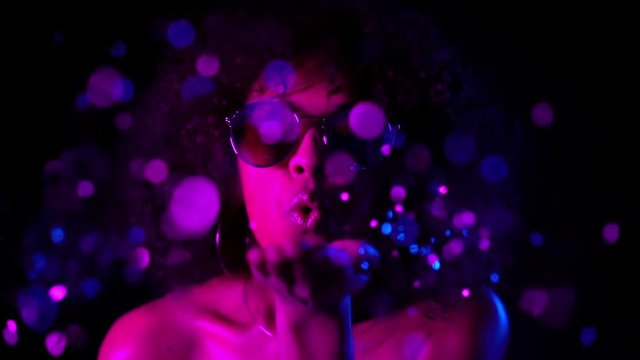 Mixed race girl with curly hair blowing confetti on dark background in neon light. Woman in glasses celebrating, depicts joy and happiness. Success, party, holiday concept