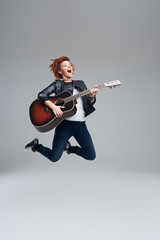 Young woman musician with an acoustic guitar in hand on a gray background. She laughs and jumps...