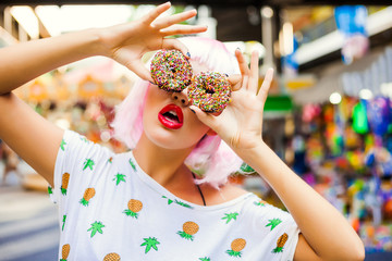bright fashion funny girl in pink wig posing on background carousel at an amusement park. The hands holding the sweet donuts