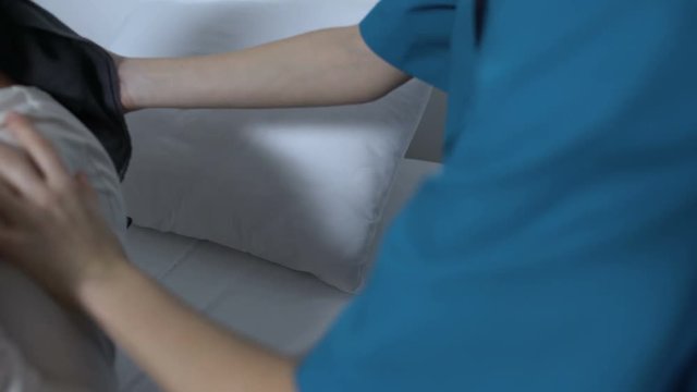 Sick-nurse helping senior patient suffering caner to lay in hospital bed, care